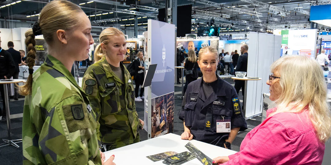 Three uniformed exhibitors speaking with a visitor at a stand at SKYDD.
