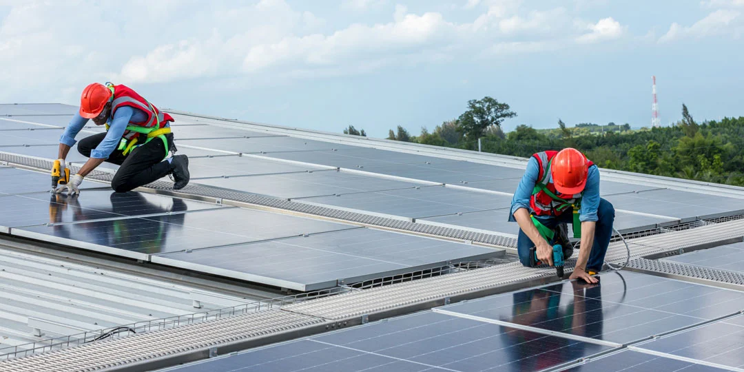 Two people wearing red helmets and safety vests installing solar panels on a roof under a clear sky.
