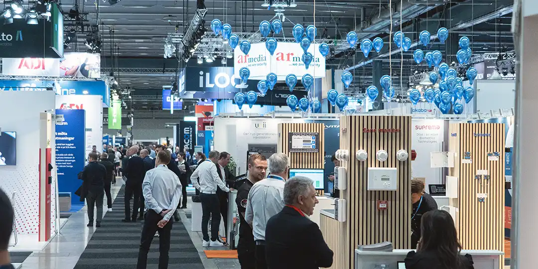 Visitors walking between stands at SKYDD in a large exhibition hall with blue balloons suspended from the ceiling.