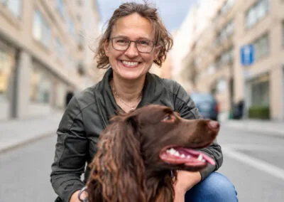 Kvinna med glasögon ler mot kameran medan hon sitter på huk bredvid en brun sökhund på en stadsgata.