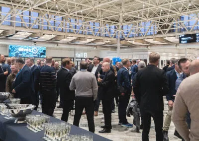En stor grupp människor minglar i en ljus mässhall, med glas och dryckesbord i förgrunden.